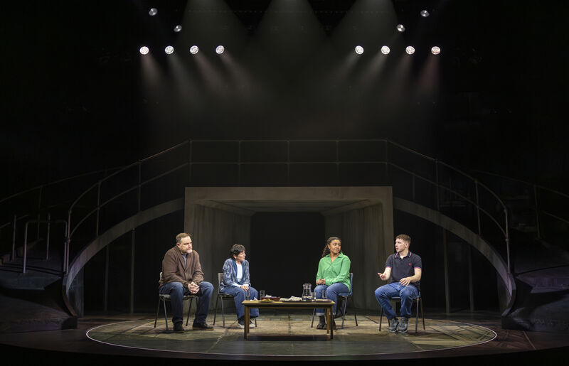 Four actors sit on a stage. Their chairs and a table are encircled by a line of light on the floor. A young man talks, his arm reaching out, whilst the others listen intently. 