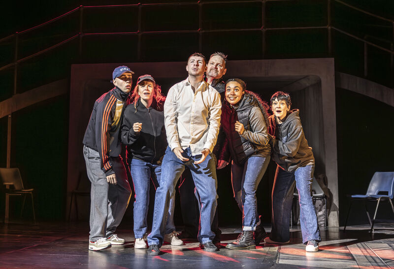 A group of 6 people wearing blue jeans and dark jackets crowd together. The man in the middle wears a white shirt. They all look up and out expectantly. They are lit with a red light. 