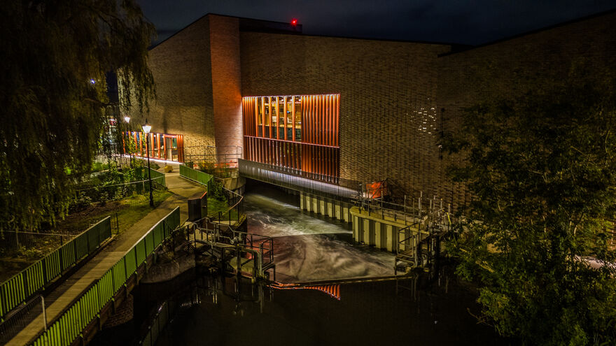 Night view image of BEAM from exterior. River flowing underneath the venue with the lights on inside.