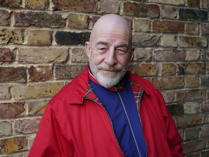 man (Jeff Innocent) wearing red jacket stood in front of brick wall