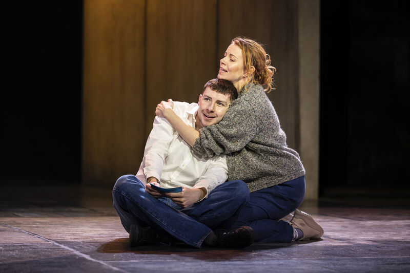 A woman with red hair kneels on the floor holding a young man in her arms. The man sits cross legged, wearing a white shirt and is resting his head on her. They both smile. 