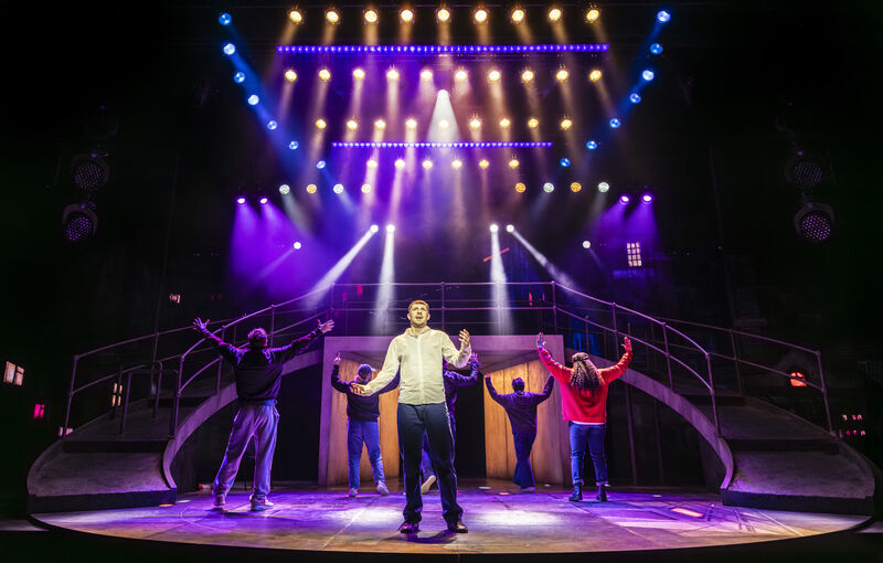A stage set with sweeping ramps down both sides and strong spotlights, in shades of blue, pink and purple. 6 actors fill the stage. All but one face the back, their arms raised. The man at the front holds his arms out, as if asking a question. 