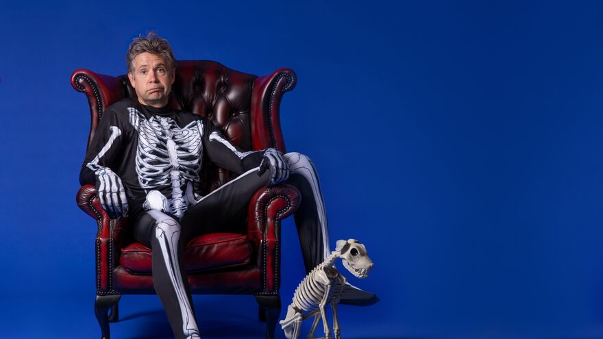 Rob Rouse sat in armchair wearing a skeleton suit, with dog skeleton sat beside him