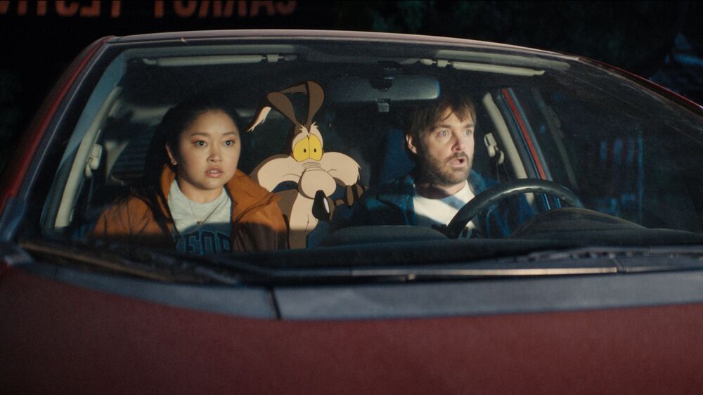 A man and a woman sit in a car with the animated Coyote character sat in the back seat