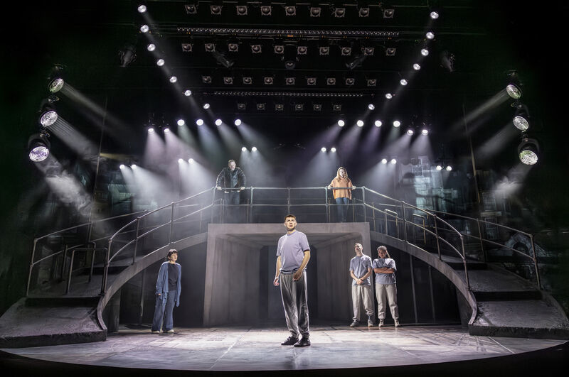A stage set with sweeping ramps down both sides and strong spotlights. 6 actors are spaced around the set, five of them look at one man in the middle, who looks out. 