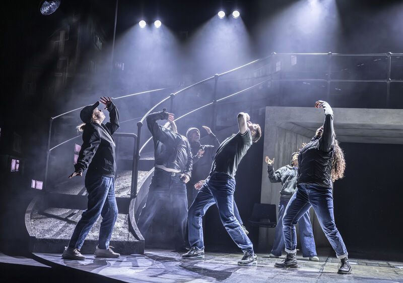 A group of 6 people in casual clothing lean back and lift an arm to their mouths, as if drinking from a cup or glass. The stage is lit with bright light and in the background, a sweeping ramp and concrete tunnel can be seen. 