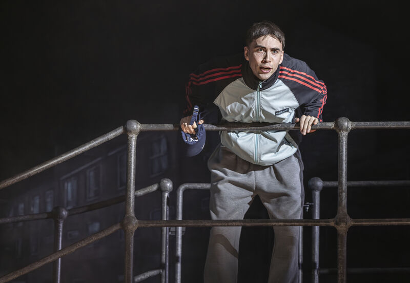 A young man with dark brown hair, wearing an adidas tracksuit top, leans forward on a steel railing. He grasps a baseball cap in his right hand. He is speaking intently to someone off camera. 