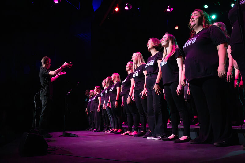A large choir sing on stage under a pink spotlight