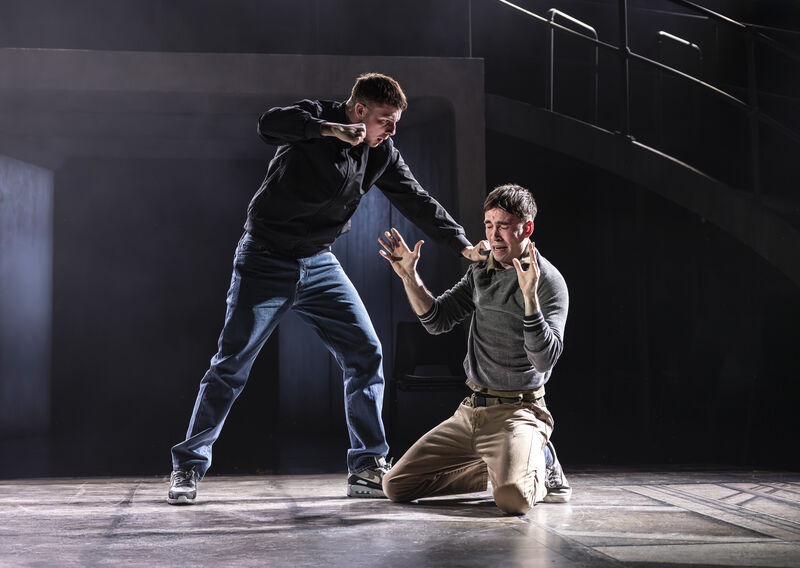 A young man in jeans and a dark jacket holds another man down by his shoulder and raises his right fist, as if about to hit him. The other man kneels on the stage, his arms raised, eyes closed and a look of fear on his face. 