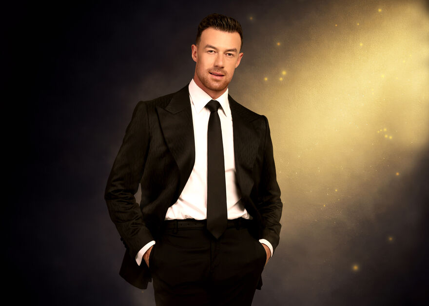 Kai Widdrington in a black suit with his hands in pockets and golden background