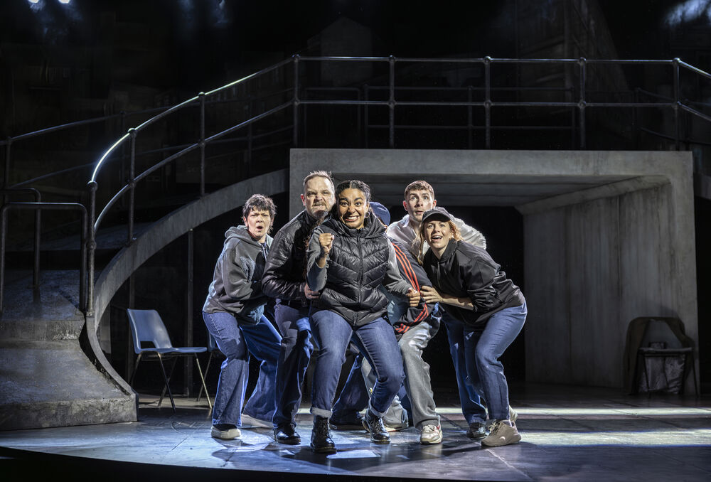 A group of 6 people wearing blue jeans and dark jackets crowd together. They look out with excited, expectant expressions. The woman at the front clenches her fist in victory. 
