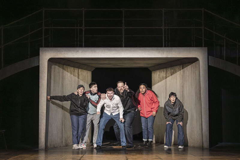 A group of 6 people in casual clothes huddle together at the end of a concrete tunnel. They look out, focussed on something in the distance. 