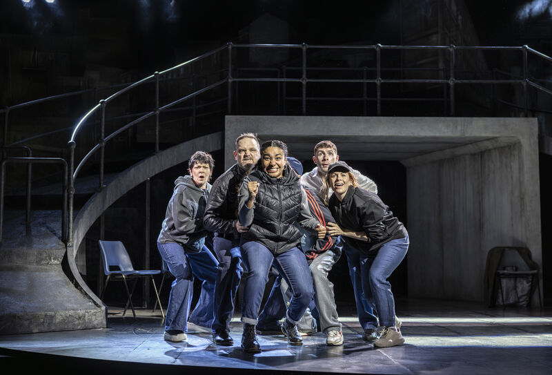 A group of 6 people wearing blue jeans and dark jackets crowd together. They look out with excited, expectant expressions. The woman at the front clenches her fist in victory. 