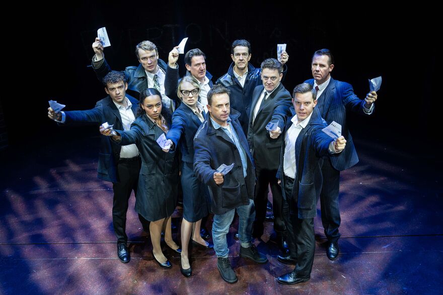 Cast huddled together each holding a dollar note up towards the camera