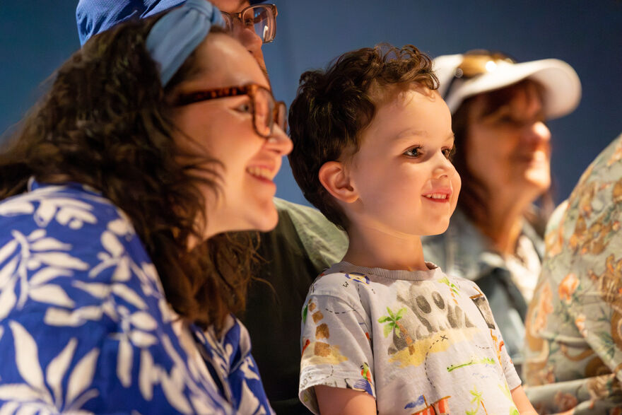 Side profile of a woman with long brown hair and glasses with a young child both looking and smiling at something out of frame.