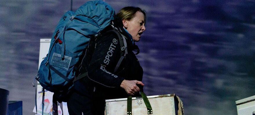 A woman walking across stage in outdoor wear with a rucksack and carrying a metal case.
