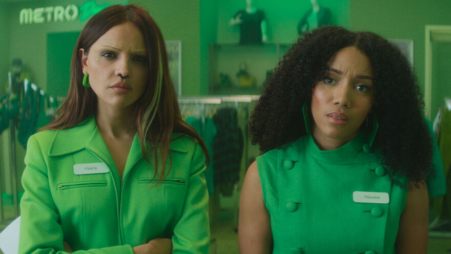 Two women sat in a green shop, wearing green uniform looking confused at the camera