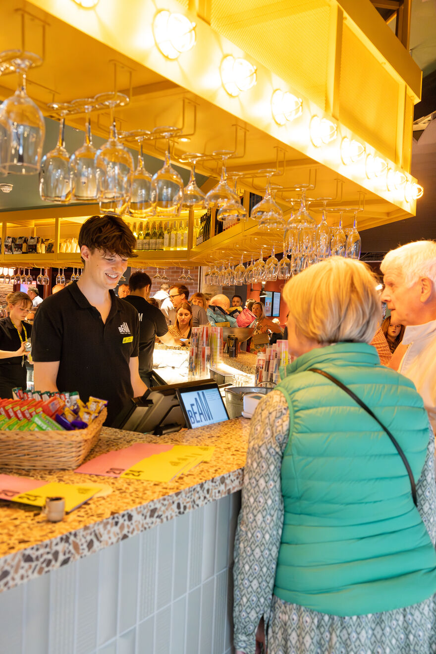 A member of the Welcome Team serves two customers from behind the bar.